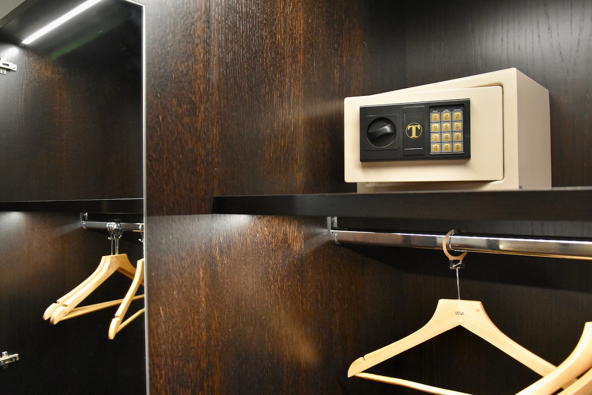 A modern wardrobe is shown, featuring dark wood paneling. A secure safe is positioned on a shelf, next to several wooden hangers. Reflective surfaces enhance the organized appearance of the storage space.