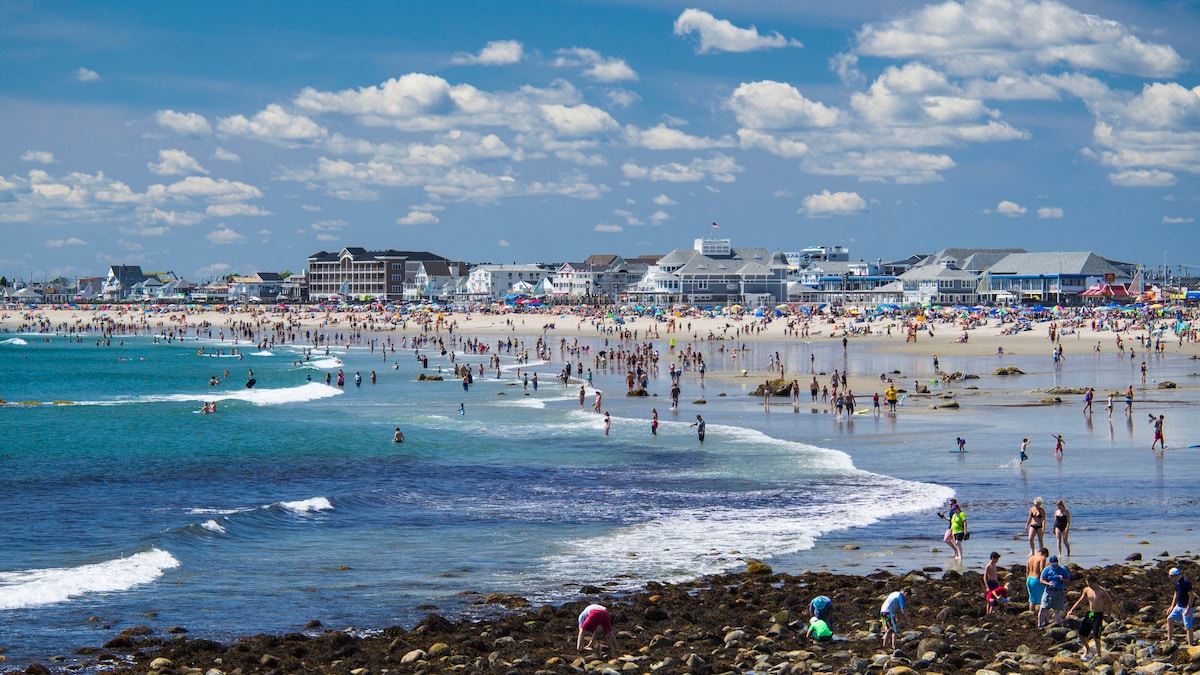 A vibrant beach scene unfolds with numerous visitors enjoying the shoreline. Waves gently lap at the sandy beach, while others explore rocky areas. Lush clouds decorate a bright blue sky, and various beachfront buildings are visible in the background, illustrating a lively coastal atmosphere.