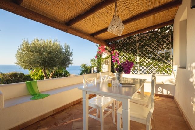 An outdoor terrace is shown, featuring a white dining table with four chairs. A decorative area is enhanced by greenery and colorful flowers climbing a trellis. The sea can be seen in the distance, with soft sunlight illuminating the space under a natural bamboo covering.