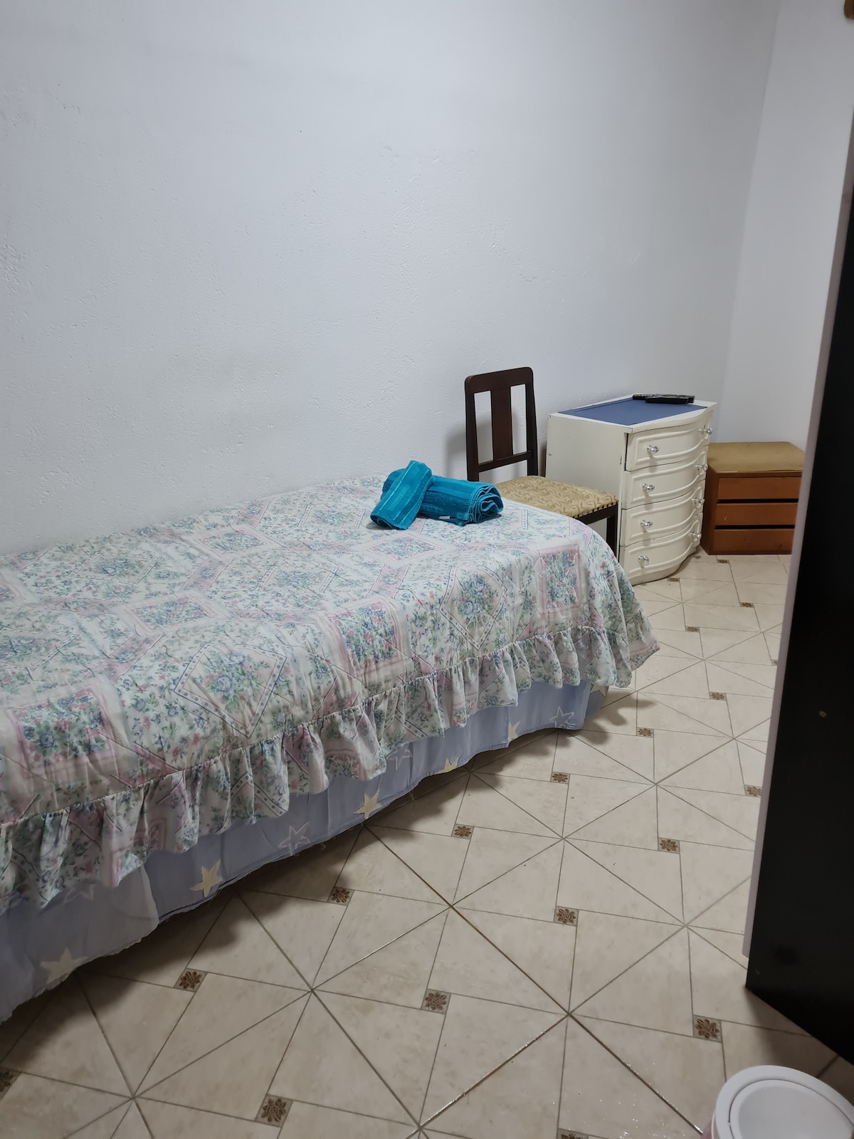 A simple bedroom features a single bed with a floral-patterned coverlet. A chair and small desk are positioned nearby, while a white dresser stands against the wall. Coordinated towels are neatly placed on the bed, with a light tile flooring completing the space.