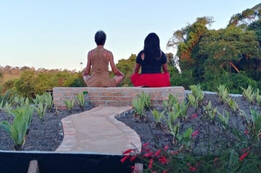 Two individuals are seated in a meditative pose on a stone platform bordered by a pathway. Surrounding greenery and flowering plants create a serene environment, with trees visible in the distance under a clear sky.