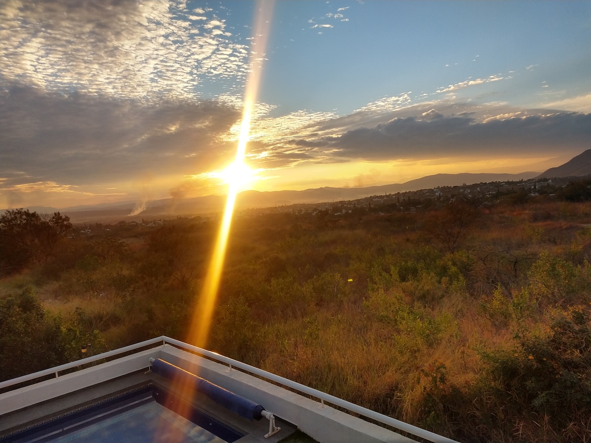 The image captures a vibrant sunset illuminating the landscape, with golden rays streaming across the sky. A portion of a heated jacuzzi is visible in the foreground, framed by lush greenery and hills in the distance, enhancing the tranquility of the setting.