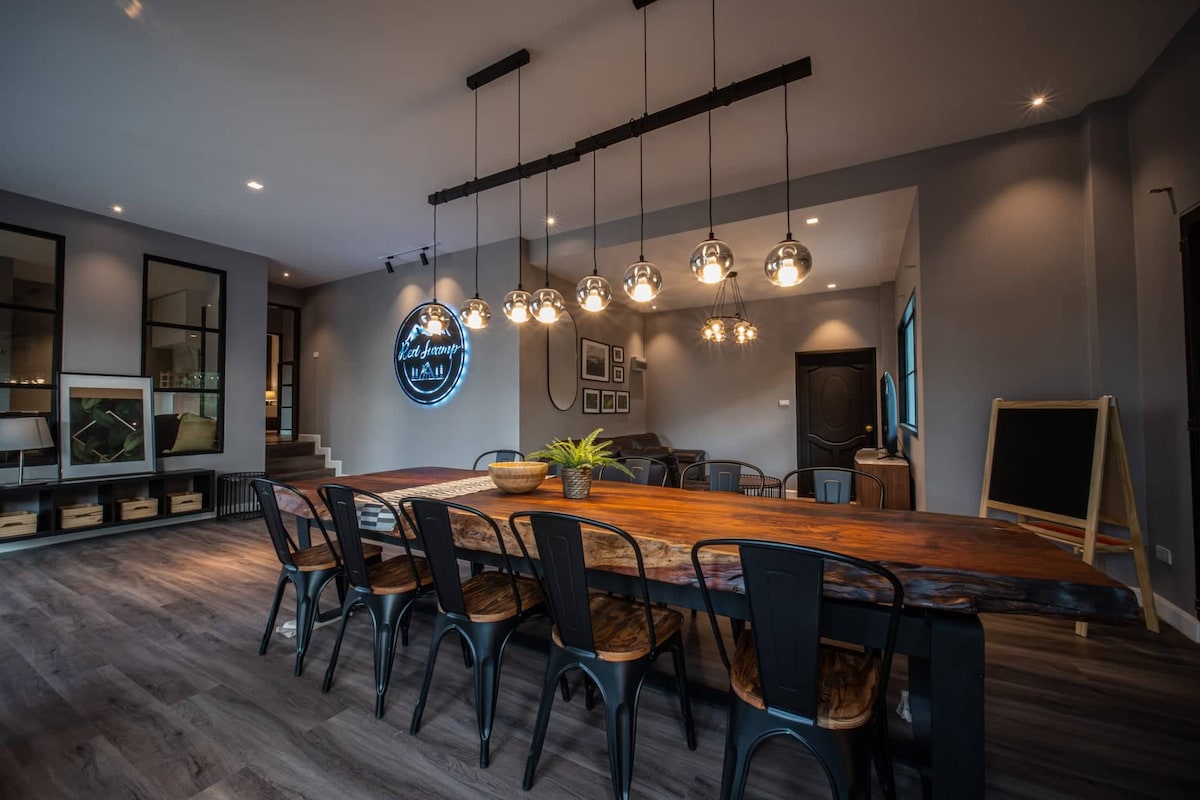 A spacious dining area features a long wooden table surrounded by numerous black chairs. Multiple pendant lights hang from the ceiling, illuminating the space. The room is decorated with framed artwork and has a cozy seating area with a sofa, enhancing the inviting ambiance.