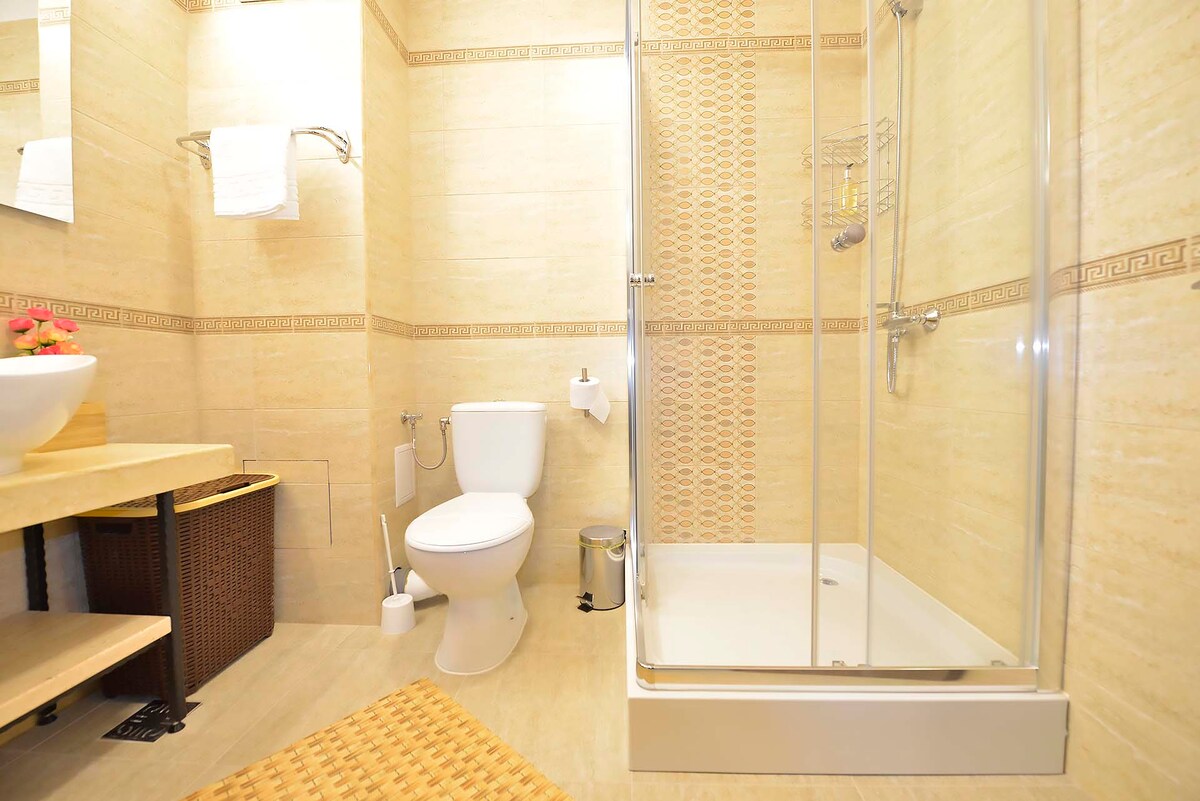 The bathroom features a glass-enclosed shower, a modern toilet, and a wooden vanity with a round sink. A woven mat covers the floor, and light tones enhance the space. Fresh towels are neatly arranged, offering a clean and welcoming environment.