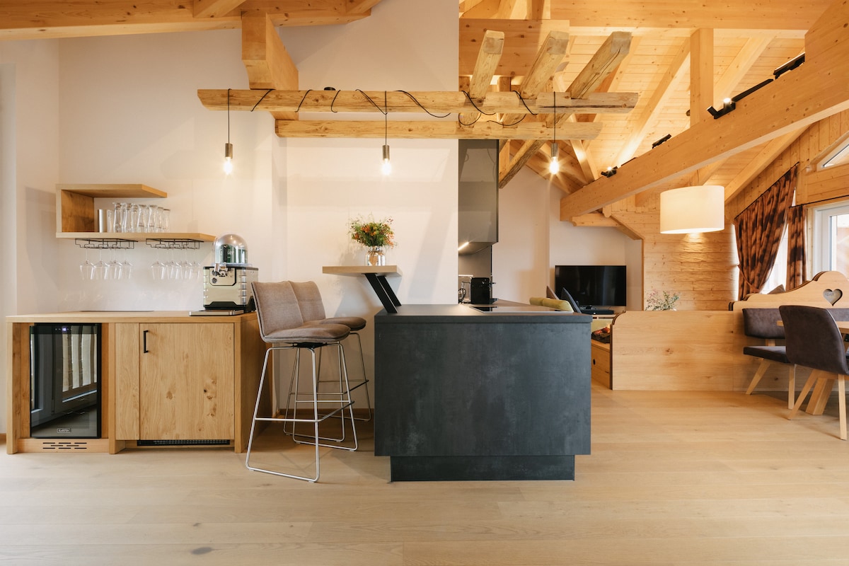 The open kitchen area is characterized by natural wood beams and a contemporary design. A central island features dark cabinetry, complemented by bar stools. An array of glassware is displayed on a nearby shelf, while natural light fills the space through large windows.