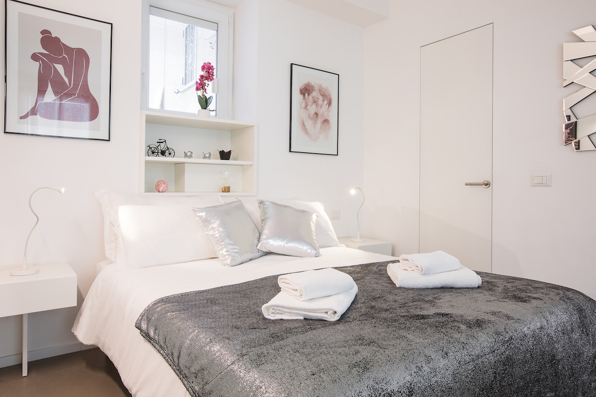 A well-lit sleeping area features a spacious bed dressed in a silver blanket, complemented by neatly arranged towels. On the wall, framed artworks add aesthetic appeal, while a shelf holds decorative items. Natural light enters through a nearby window.