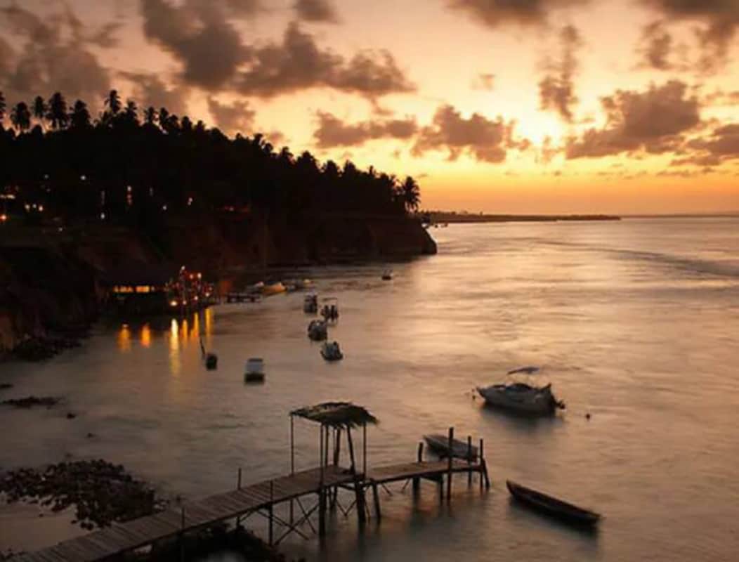 A serene waterfront is depicted at sunset, with soft hues of orange and pink reflected on the water's surface. Small boats are anchored in the bay, and a wooden pier extends into the water, surrounded by lush vegetation along the shore.