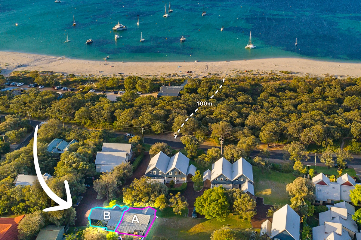An aerial view highlights the cottage's location within a leafy garden setting. The image shows proximity to the calm waters of Geographe Bay, approximately 100 meters away, with boats gently anchored nearby and sandy beach strips visible along the shoreline.