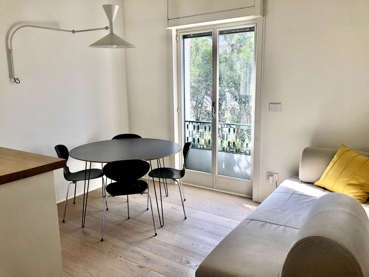 A bright living area features a round dining table surrounded by four black chairs. A light-colored sofa with a yellow cushion is positioned nearby. Large glass doors open to a balcony with views of greenery.