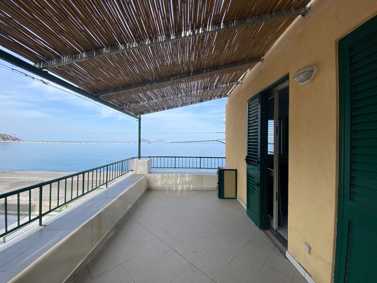 A spacious terrace offers a relaxing outdoor space with a shaded roof overhead. It features a tiled floor and a low wall, providing a clear view of the nearby beach and sea. The entrance to the interior is visible, framed by green shutters.
