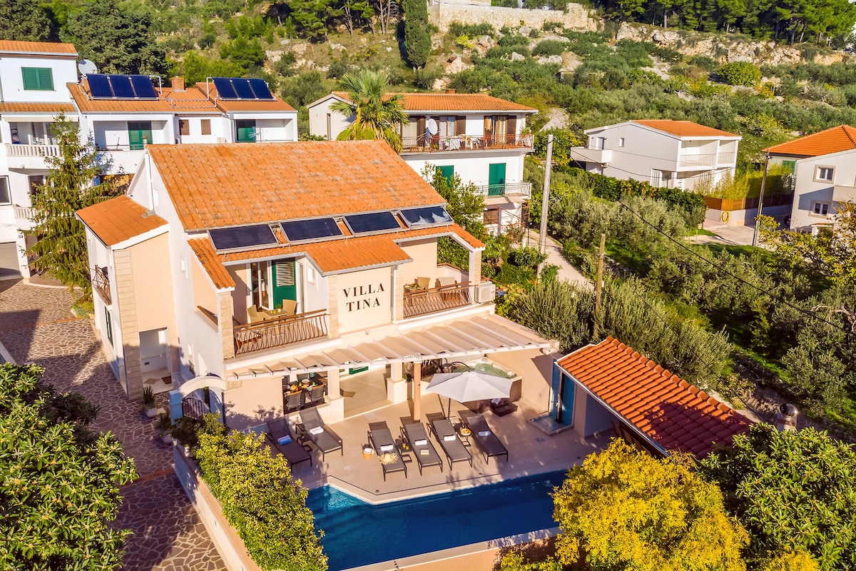 An aerial view of Villa Tina is displayed, featuring its terracotta roof and spacious outdoor area. The private heated pool is visible, surrounded by sun loungers, while the landscaped gardens and nearby residential buildings create a serene setting.