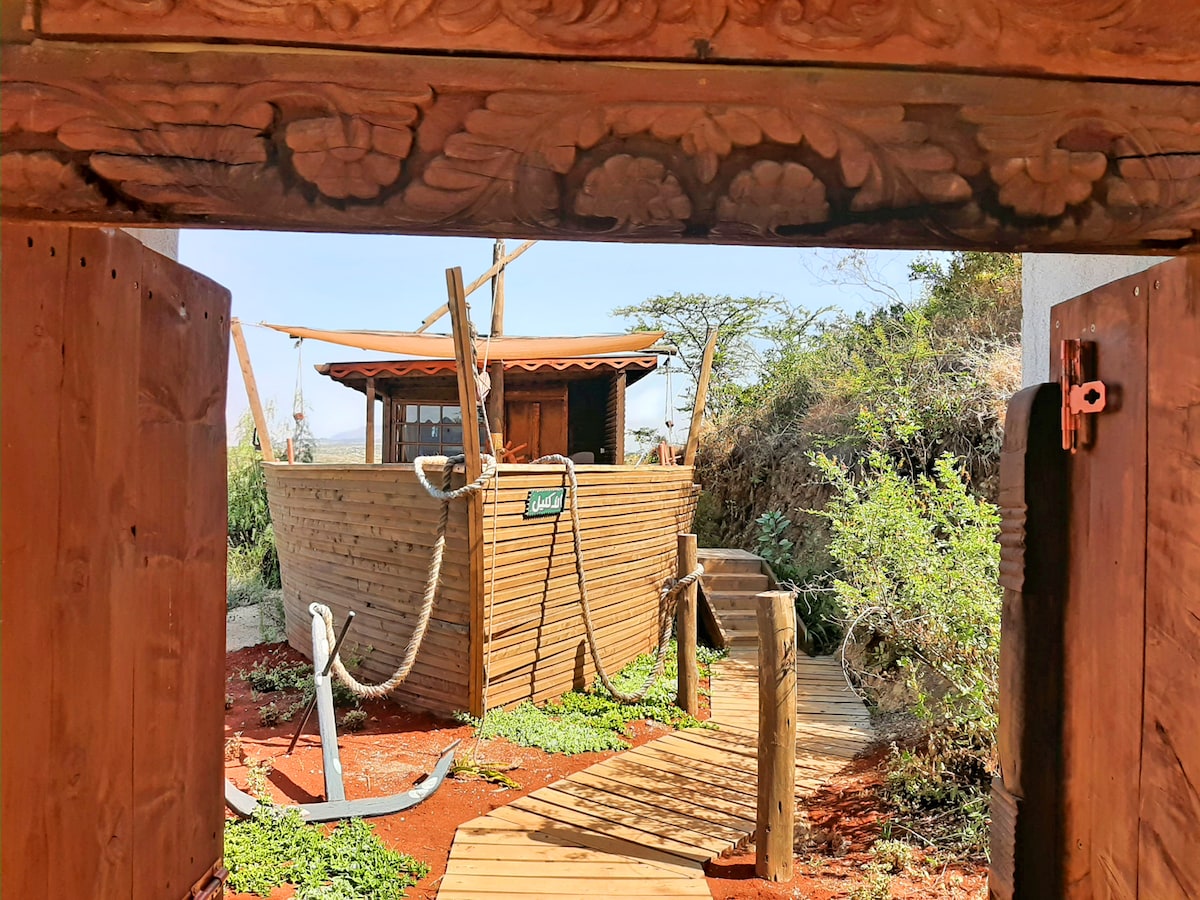 An entrance framed by intricately carved wooden doors leads to a wooden structure. The path is lined with greenery, bordered by rustic wooden decking. Sunlight illuminates the scene, highlighting the anchor positioned near the entrance.