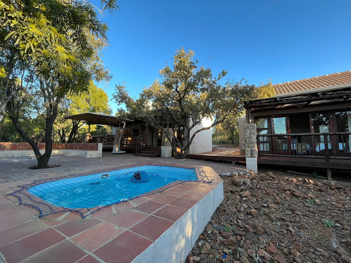 A private swimming pool is set within a tiled area, surrounded by a natural landscape. The house features a spacious deck with comfortable outdoor seating under a shaded structure, complemented by trees providing greenery. The clear sky enhances the peaceful outdoor setting.