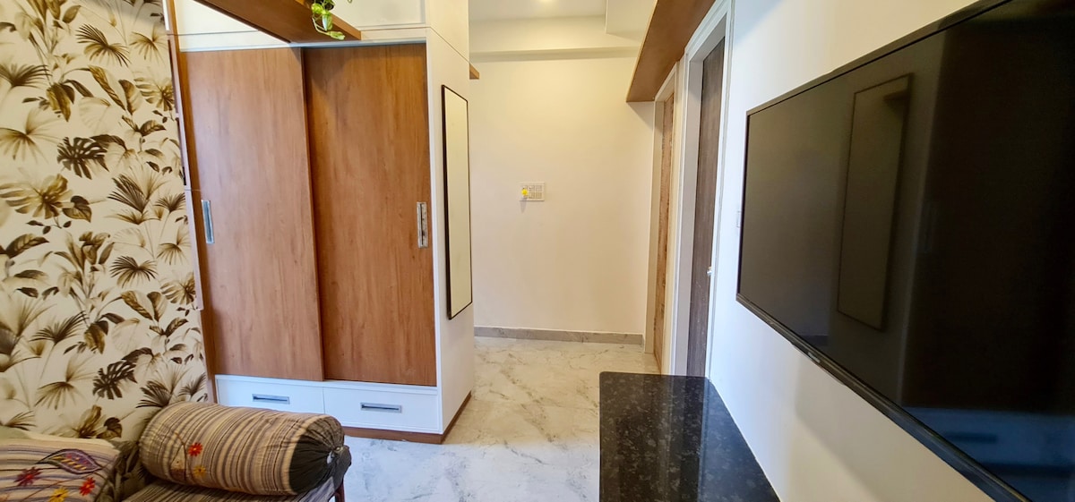 A spacious living area showcases a comfortable sofa with patterned upholstery, a mounted television, and sleek wooden wardrobes. The light marble flooring adds a contemporary touch, while neutral walls contribute to a calming ambiance.