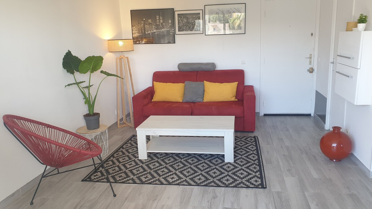 A contemporary living area features a red sofa adorned with yellow cushions, complemented by a white coffee table on a patterned rug. A large plant is displayed in a pot next to a standing lamp, and an accent chair adds a pop of color to the space.
