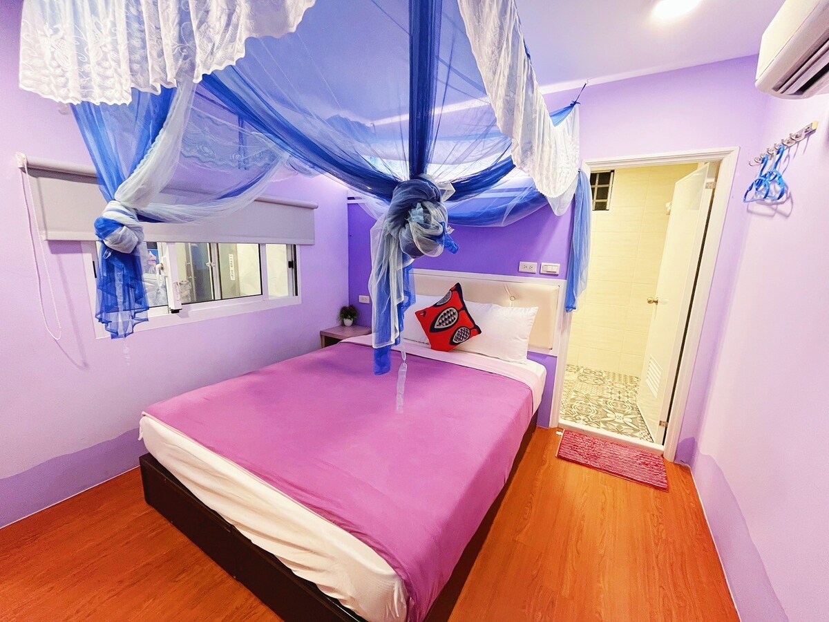 A bright bedroom features a purple color scheme with a spacious bed covered in light bedding. Sheer blue drapery hangs from the ceiling, adding a soft touch. A small plant decorates the bedside, and a private bathroom can be seen through an adjacent door.