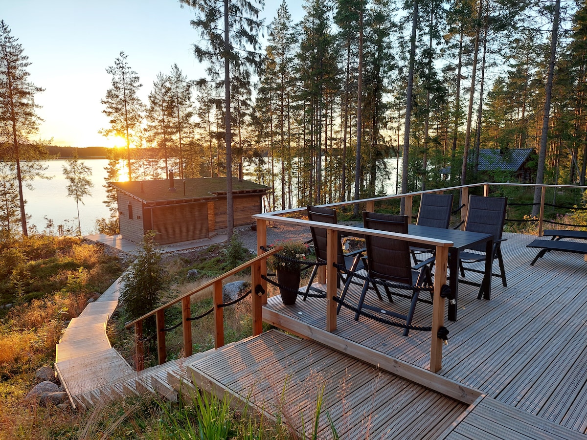 An outdoor terrace features several black chairs arranged around a wooden table, overlooking a tranquil lake at sunset. The surrounding landscape includes trees and a sauna building in the background, with the warm sunlight casting a soft glow on the deck.