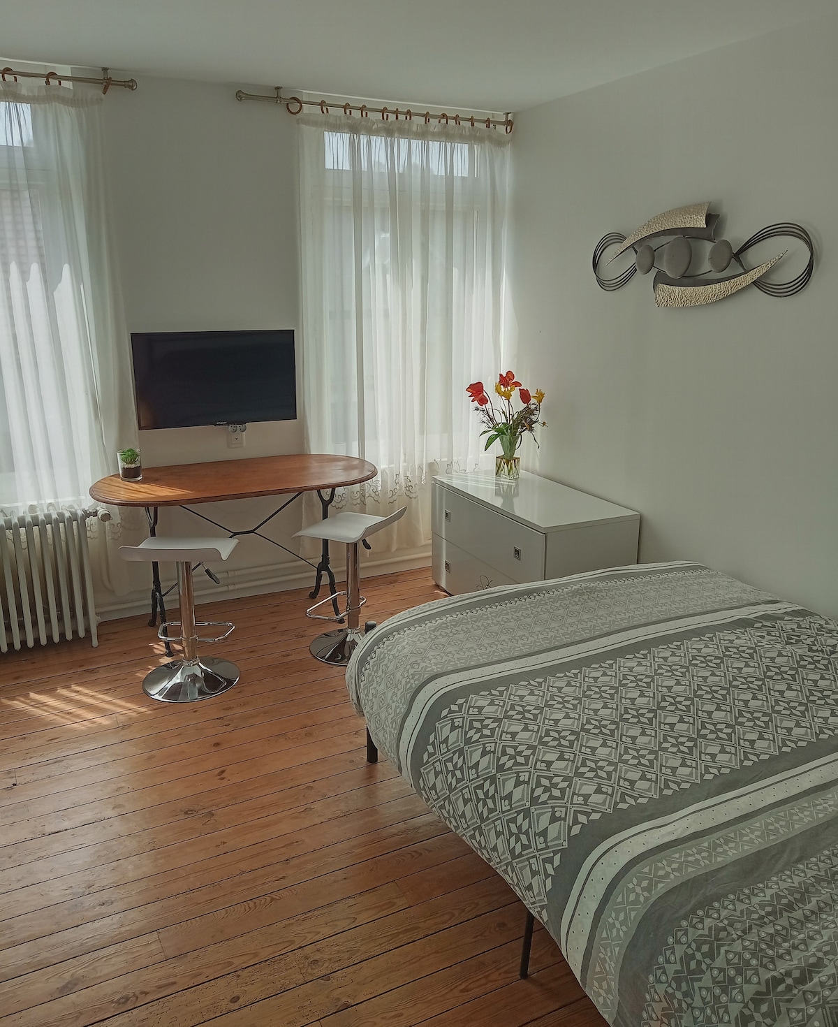 A bright studio space features a spacious bed with a patterned duvet. A small table with two stools is positioned near a window adorned with sheer curtains, allowing natural light to fill the room. A television is mounted on the wall, and a vase of flowers adds a touch of color.