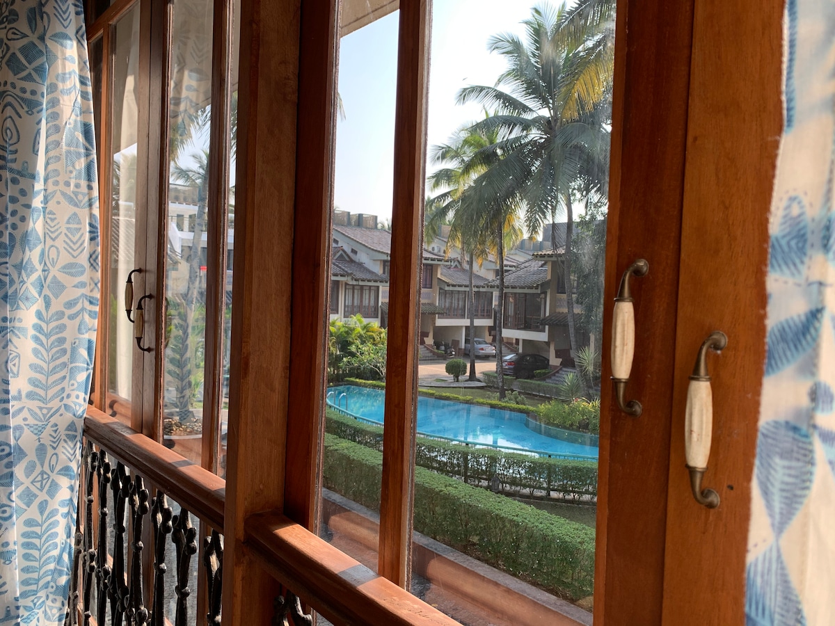 Large wooden windows offer a view of the manicured gardens and a clear blue pool. Palm trees sway gently outside, providing a refreshing atmosphere. Light fabric curtains frame the window, allowing for natural light to brighten the indoor space.