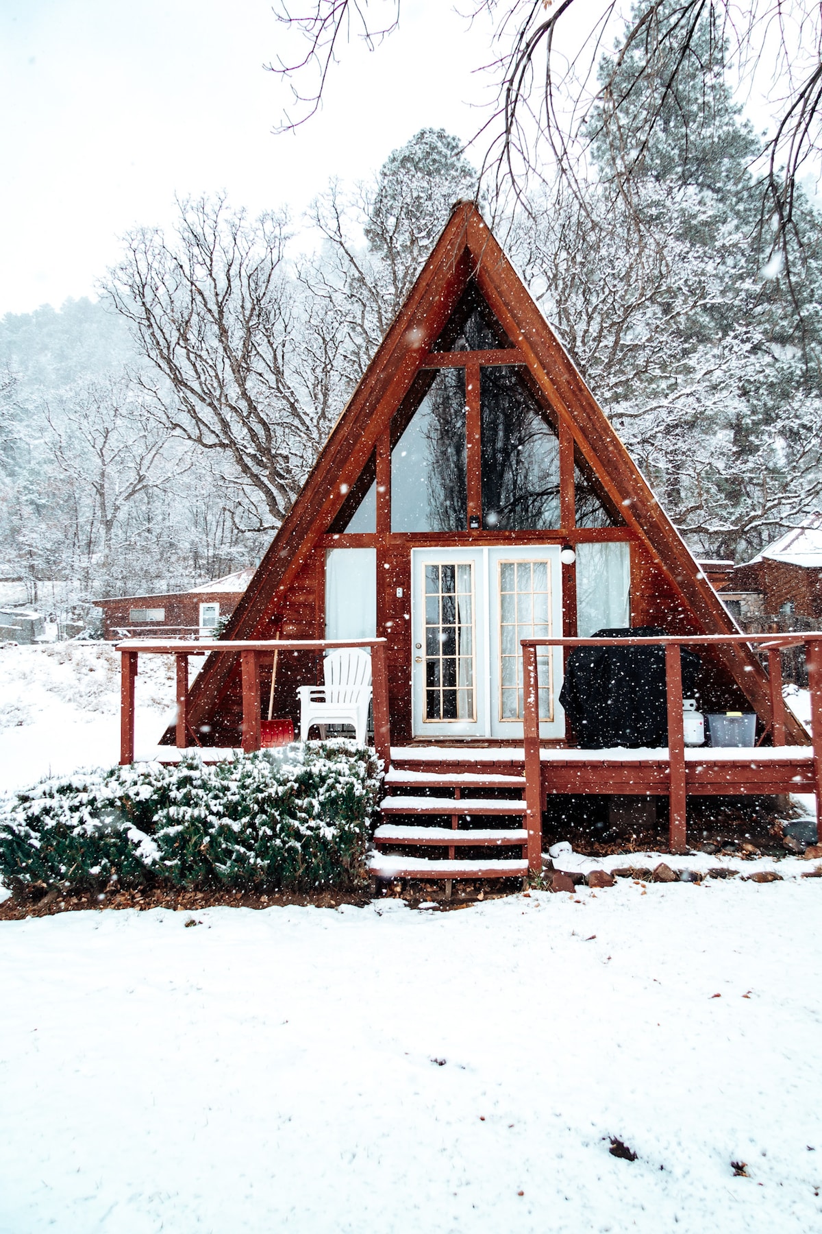 An A-frame chalet is blanketed in fresh snow, with large windows allowing natural light to fill the interior. A wooden deck is adorned with a white chair, and a lush green shrub contrasts with the snowy landscape.