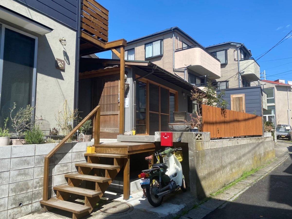 The exterior of a renovated one-story building is visible, featuring a wooden entrance with steps leading to a welcoming porch. The surrounding area is calm, with nearby modern residential structures and greenery, contributing to a tranquil atmosphere.