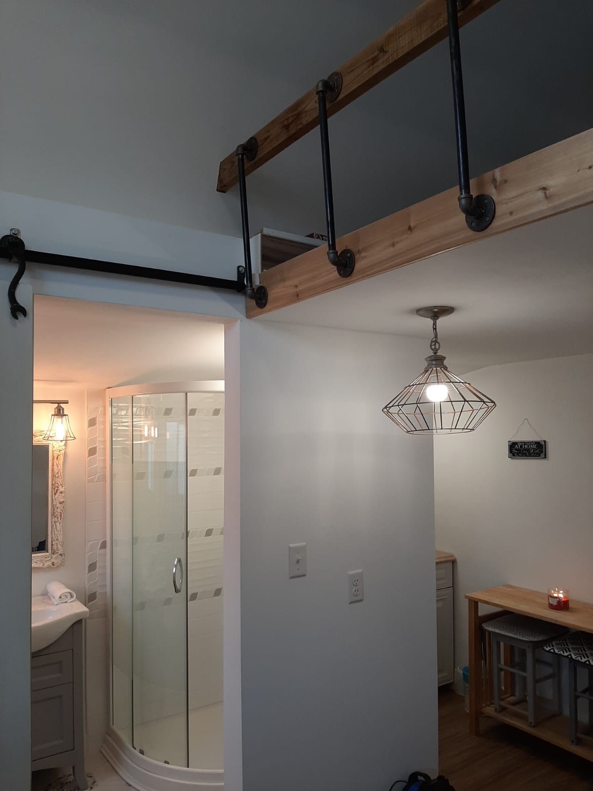 A modern bathroom is visible, featuring a glass shower stall and a vanity with a large mirror. Industrial-style lighting hangs from the ceiling, while a simple wooden table occupies the corner, adding functionality to the space.