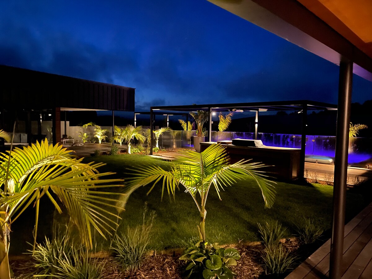 Illuminated landscape features tropical plants surrounding a modern outdoor area at night. A sleek pool reflects purple light, while the sky transitions between deep blue and gray, creating a serene ambiance. The design integrates natural elements with contemporary architecture.