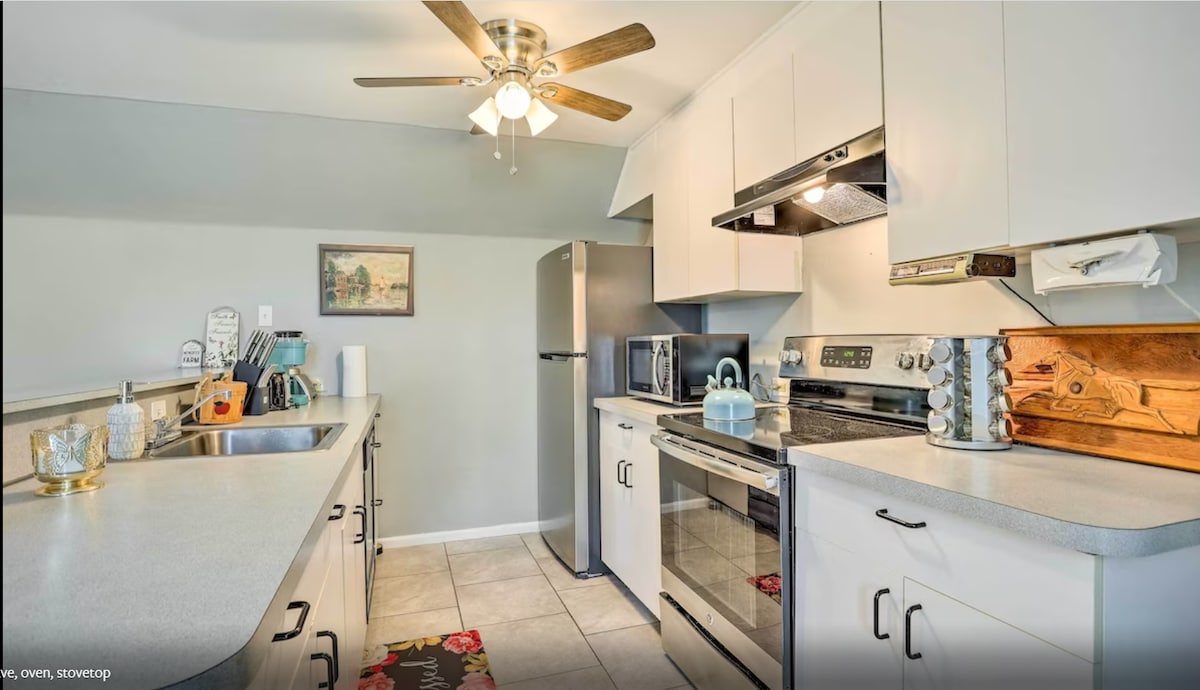The kitchen area features modern stainless steel appliances, including a refrigerator and oven. A ceiling fan provides airflow, and the countertops are crafted from smooth, light-colored materials. Overhead cabinets offer storage, while decorative items add a touch of warmth to the functional space.