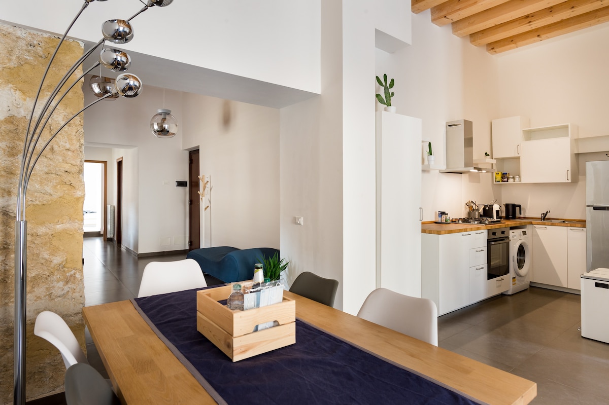 The spacious dining area features a wooden table with a blue runner, surrounded by modern chairs. A sleek kitchen is seen in the background, equipped with essential appliances. Natural light filters through the open layout, highlighting the warm tones of the wooden beams above.
