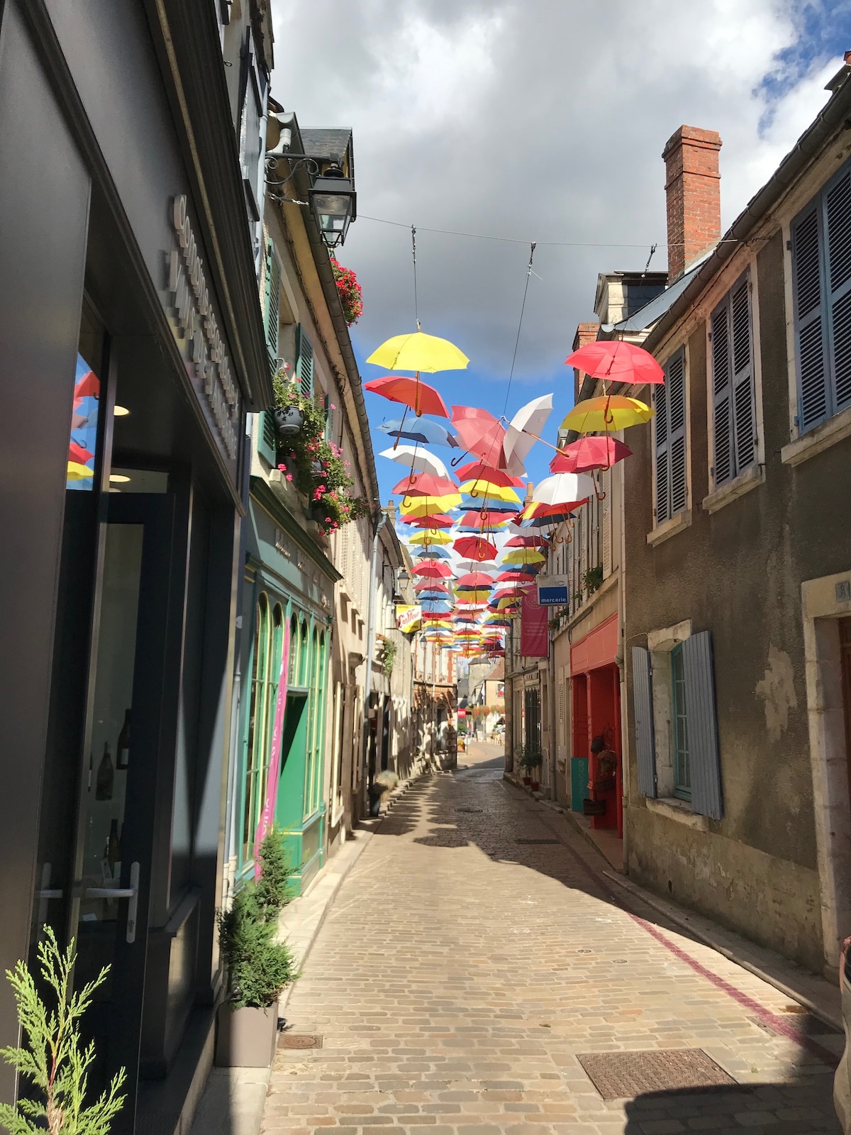 A charming street is adorned with colorful umbrellas suspended overhead, casting playful shadows on the cobblestone pathway. Traditional buildings line both sides, showcasing a mix of vibrant colors and rustic textures, contributing to a delightful atmosphere ideal for leisurely strolls.