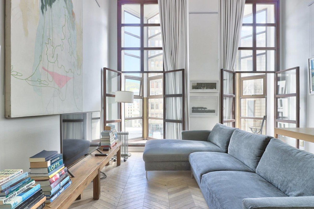 A comfortable living area features a spacious sectional sofa, complemented by a wooden coffee table stacked with books. Large windows with open panels allow natural light to fill the room, showcasing abstract wall art and a modern aesthetic.