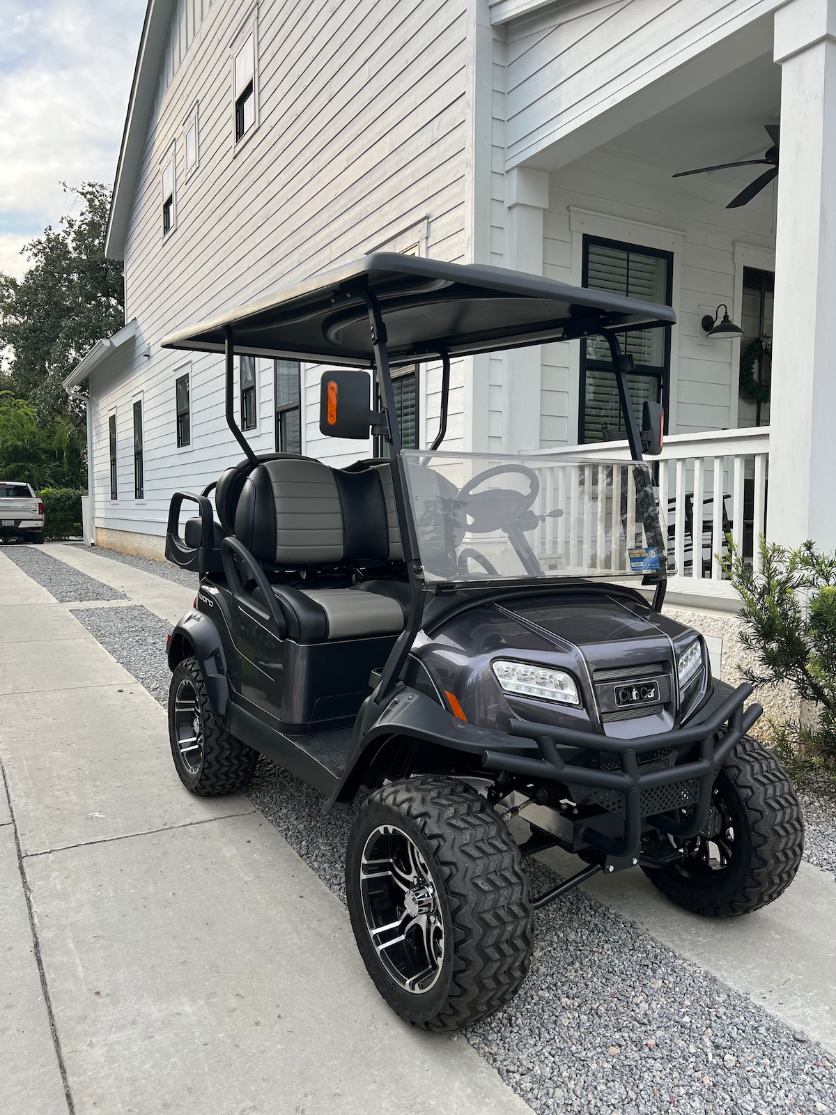 A four-seater golf cart is parked on a concrete driveway alongside a cottage. The cart features a black exterior with chrome accents and large tires. A spacious canopy provides shade, and the background includes a white house with a porch and greenery.