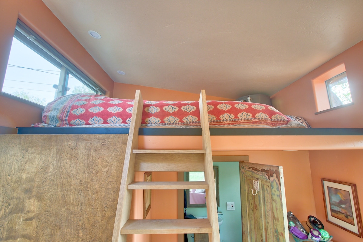 A lofted sleeping area features a spacious mattress with a decorative blanket. Natural light enters through multiple windows, illuminating warm orange walls. A wooden ladder provides access to the loft, while details of the inviting interior can be glimpsed below.