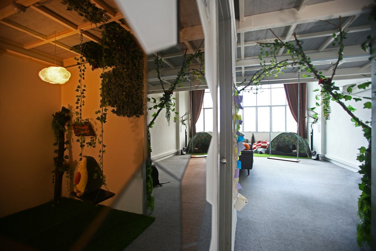 The image captures a spacious living area adorned with natural elements, featuring plush seating and two colorful tents. Soft light filters through large windows, illuminating the inviting atmosphere created by hanging greenery and decorative accents.