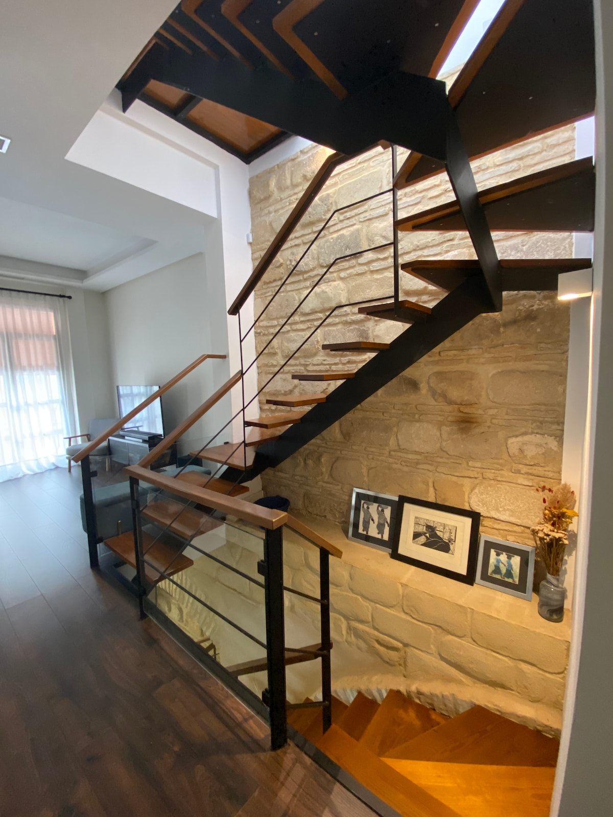 A modern staircase is showcased, featuring wooden steps and black metal railings. Natural light filters through a nearby window, illuminating the textured stone wall beside the stairs. Framed photographs are displayed on the wall, adding a touch of character to the space.