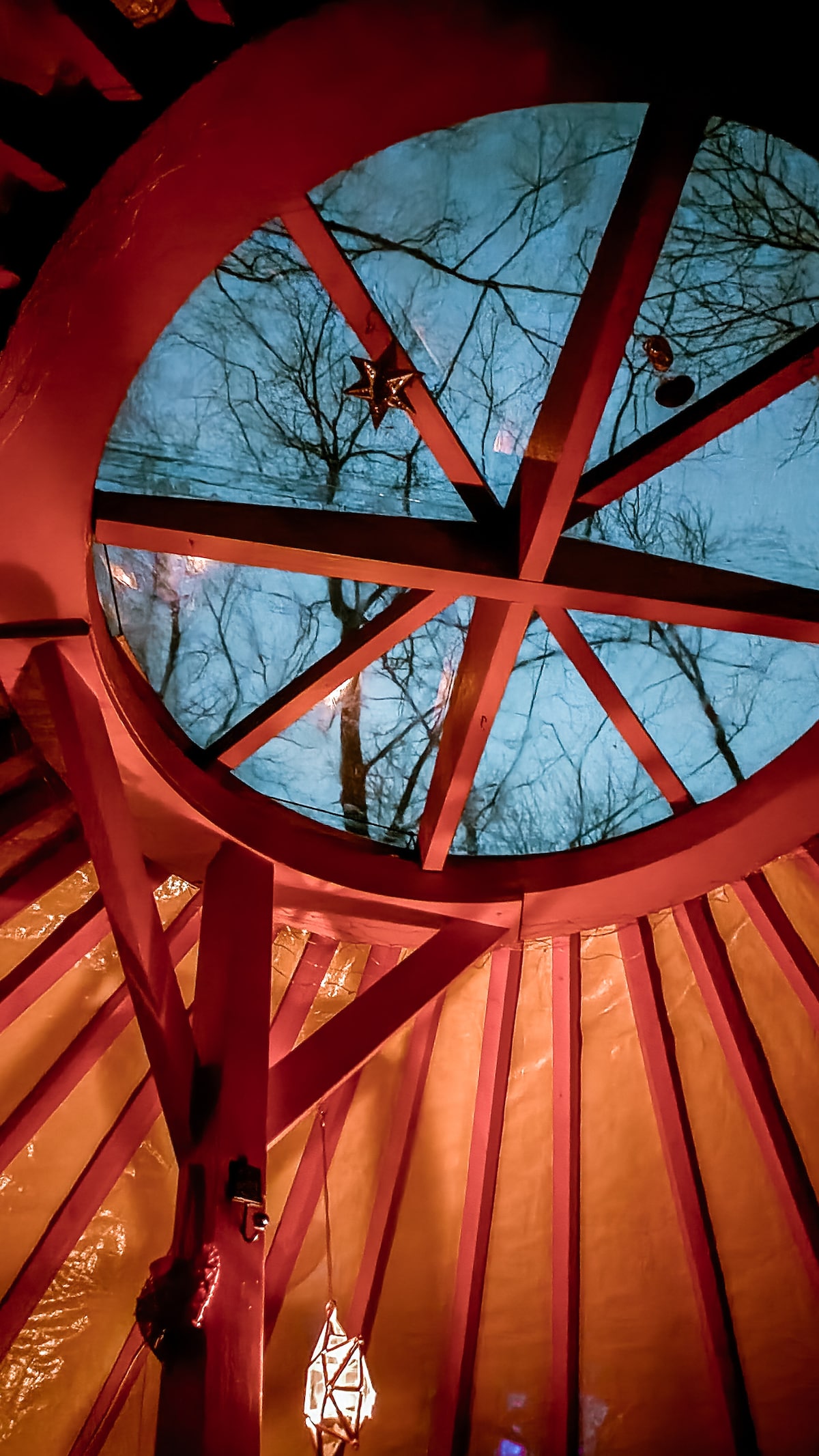 A circular skylight offers a view of the night sky framed by wooden beams. The area is softly illuminated by a hanging lantern, casting gentle light on the colorful walls. The trees outside are visible through the clear glass, enhancing the connection with nature.