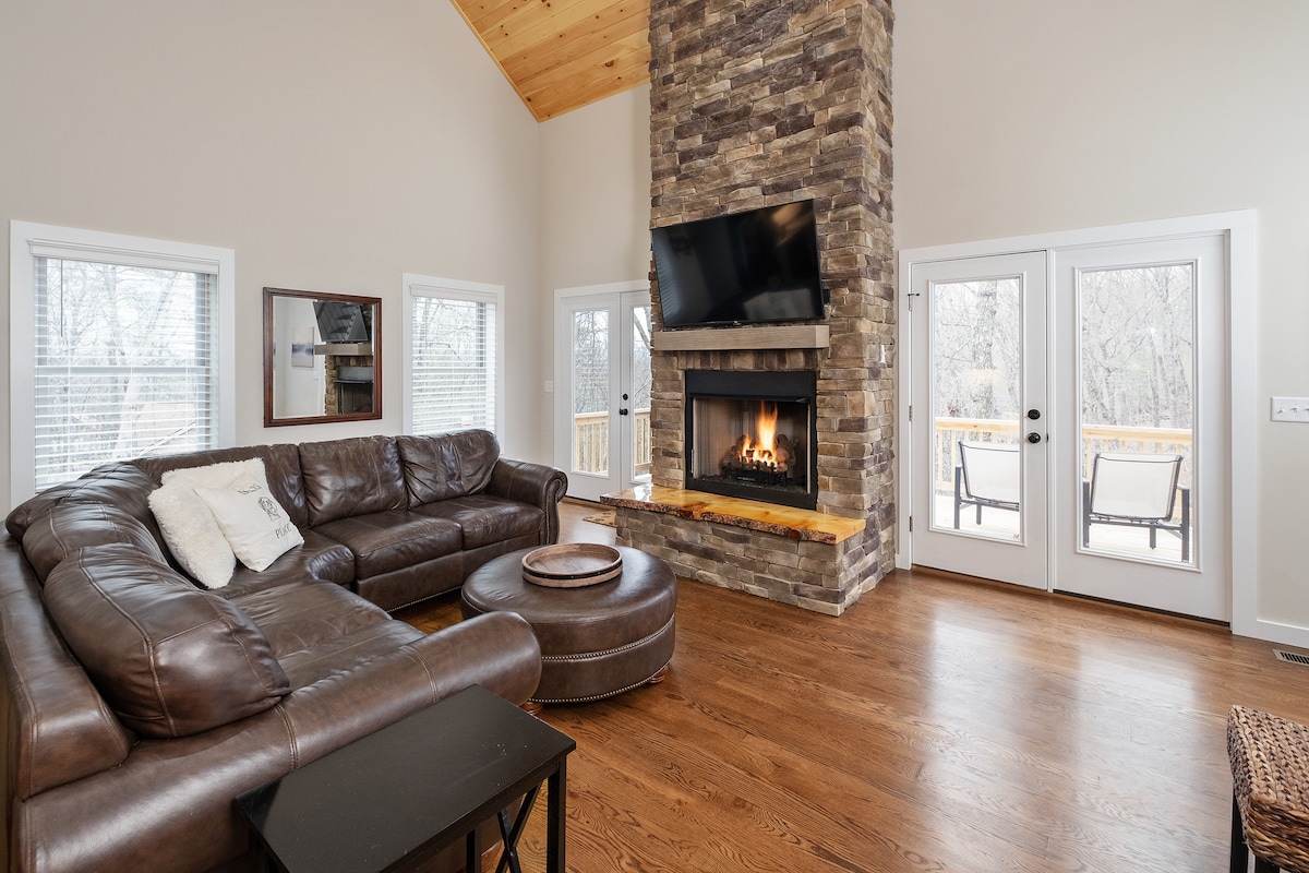 A welcoming living area features a spacious leather sectional sofa and a circular coffee table. A stone fireplace is the focal point, complemented by a flat-screen TV above it. Large windows allow natural light to fill the space, with access to a deck through double doors.