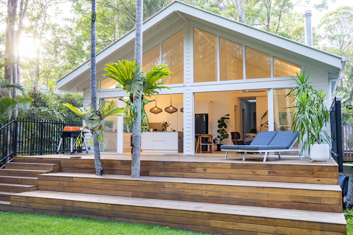 The house features large glass doors that connect the open living area to a spacious wooden deck, surrounded by lush greenery. Modern outdoor seating and tropical plants are visible, enhancing the serene atmosphere. Natural light floods the interior spaces, emphasizing the welcoming design.