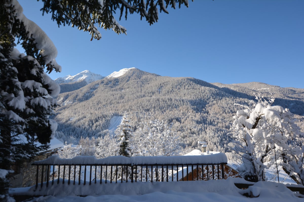 A scenic winter view is captured from the balcony, showcasing snow-covered mountains and forests. The wooden railing is blanketed in snow, and trees with frosted branches frame the image, illustrating a peaceful, natural landscape.