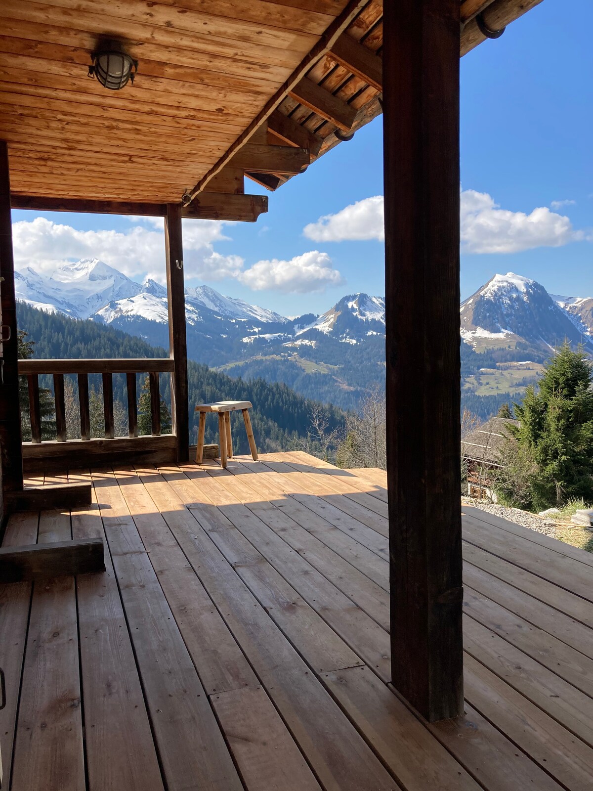 A wooden terrace with a smooth, warm-toned floor offers expansive views of snow-capped mountains and lush valleys. A sturdy wooden stool is positioned near the edge, inviting relaxed contemplation of the stunning landscape under a clear blue sky.