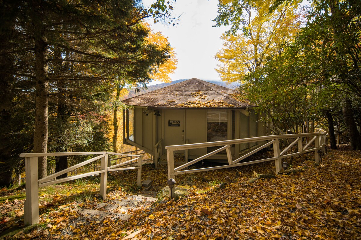 A roundhouse is positioned among autumn foliage, with leaves of orange and yellow blanketing the ground. The exterior features large windows and a sloped roof, framed by trees. A wooden railing guides the path to the entrance, enhancing the home's natural surroundings.