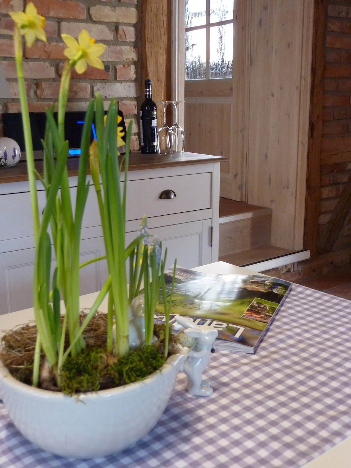 A decorative planter with green sprouts and blooming yellow flowers is placed on a checked tablecloth. A magazine is positioned nearby, alongside a bottle of wine and glassware on a white cabinet. A staircase is visible in the background, leading to an inviting wooden door.