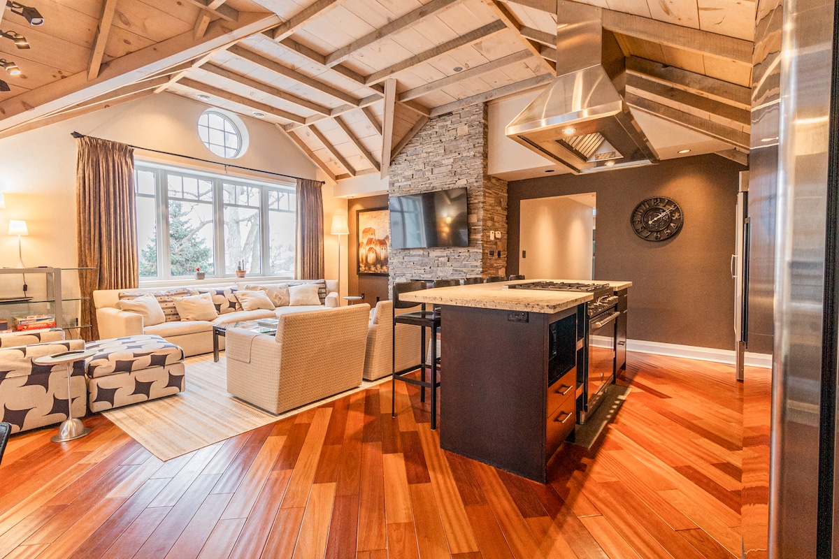An open living area features vaulted wooden ceilings and a stone fireplace. A modern kitchen with stainless steel appliances includes a commercial-grade gas stove. Large windows provide ample natural light, while warm wood flooring complements the cozy seating arrangement.