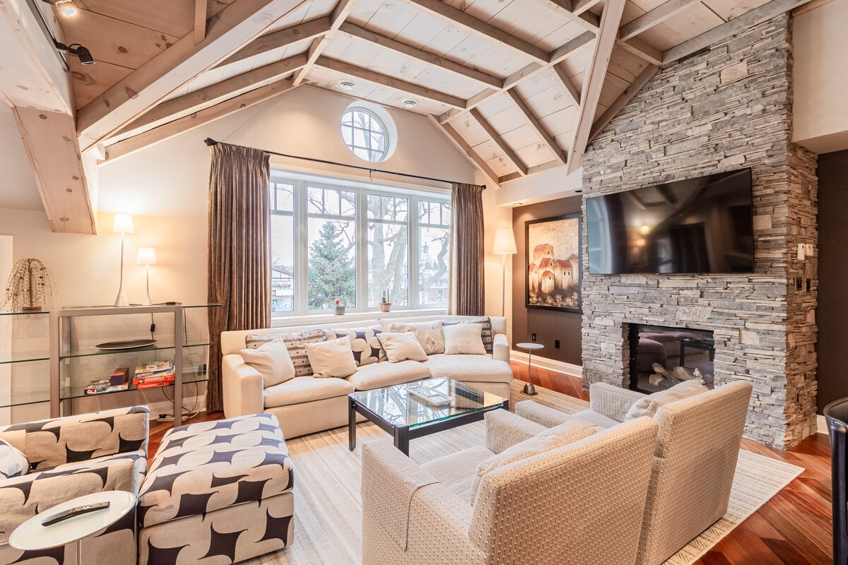 The spacious living area features vaulted wooden ceilings and large windows allowing natural light. A cozy seating arrangement includes multiple sofas and chairs arranged around a glass coffee table, complemented by a stone fireplace and wall-mounted television.