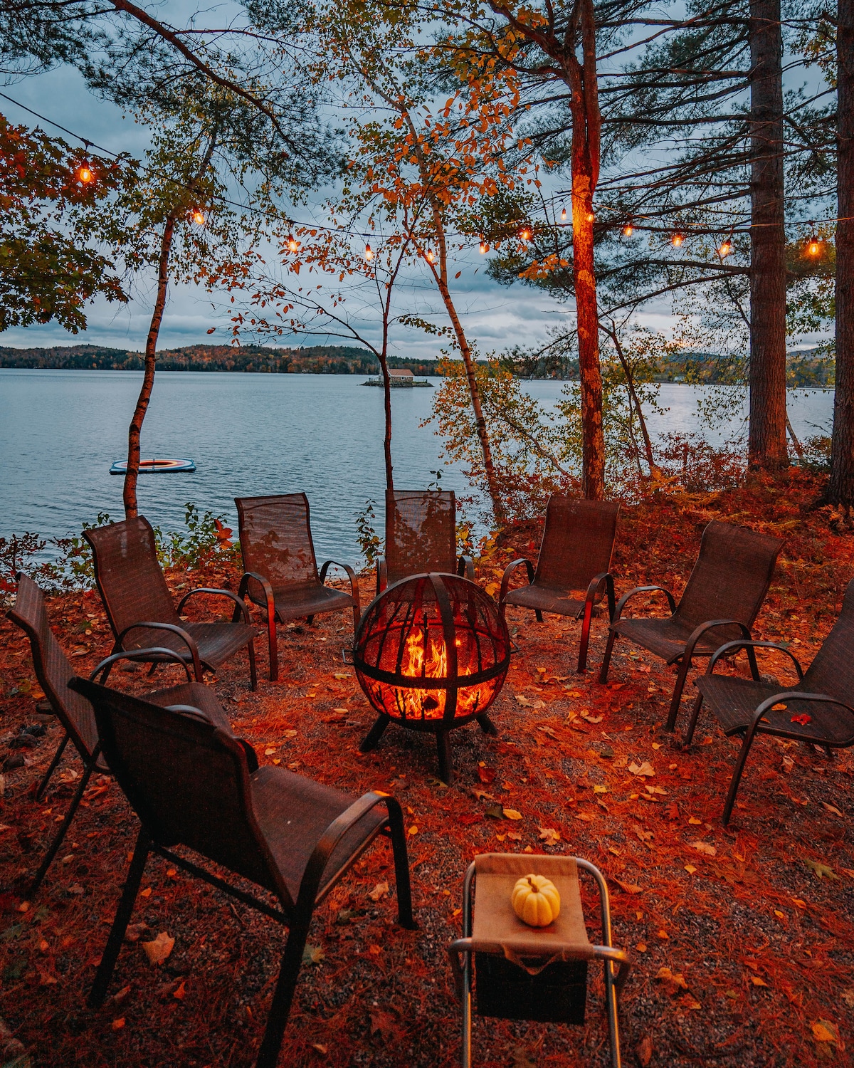 A circular arrangement of chairs surrounds a glowing fire pit, set against the backdrop of a calm lake. Autumn leaves scatter the ground, while overhead string lights softly illuminate the space. Trees stand tall on the periphery, enhancing the peaceful lakeside atmosphere.