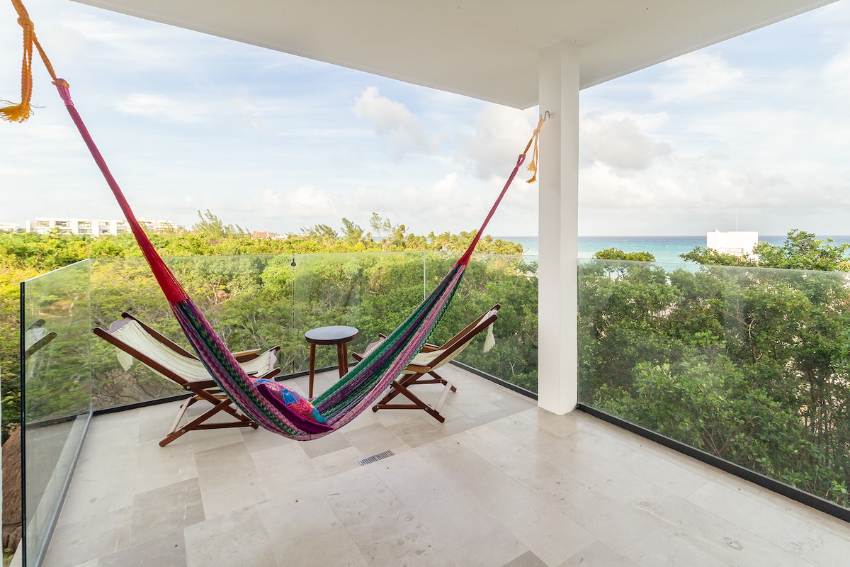 A spacious terrace is populated with a colorful hammock and two wooden chairs. A small round table sits between them. The view encompasses lush foliage leading to the ocean, under a clear sky filled with soft clouds.