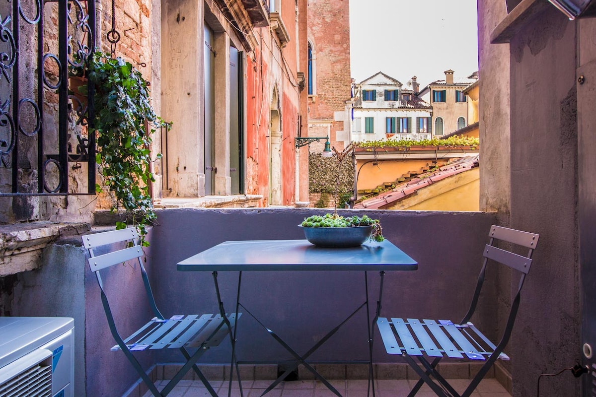 A private terrace showcases a small dining table accompanied by two metal chairs. A decorative bowl rests at the center of the table, surrounded by views of terracotta rooftops and historic buildings. Vines climb the walls, adding a touch of greenery.