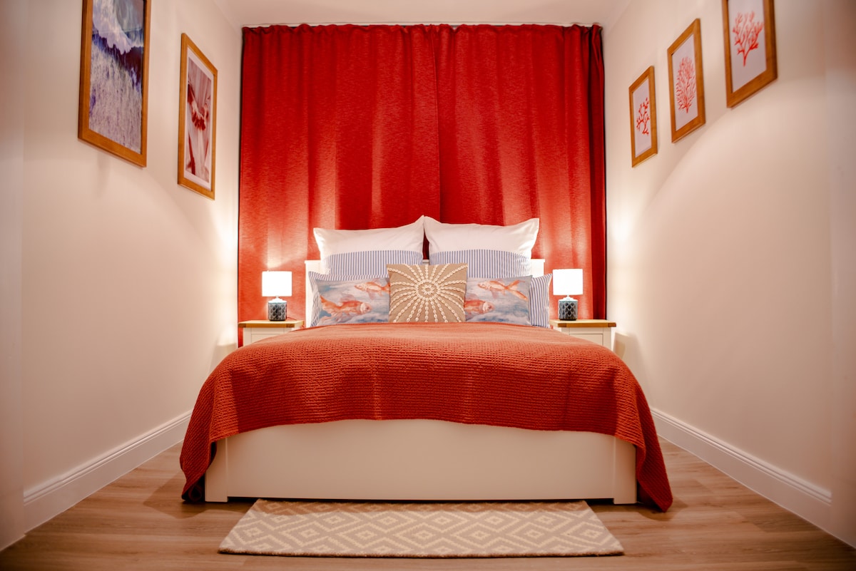 A cozy bedroom features a spacious bed with crisp white pillows and a decorative throw. Soft lighting from bedside lamps illuminates the room, while red curtains create a warm backdrop. Framed artwork adorns the walls, adding subtle colour to the ambiance.