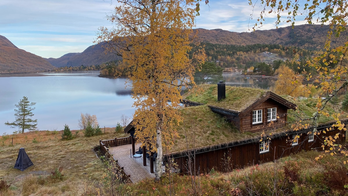 A rustic cabin is set against a serene lake, surrounded by autumn foliage. The structure features a grassy roof and wooden exterior. A fire pit is visible on a spacious patio, enhancing the natural surroundings that reflect the tranquil scenery.