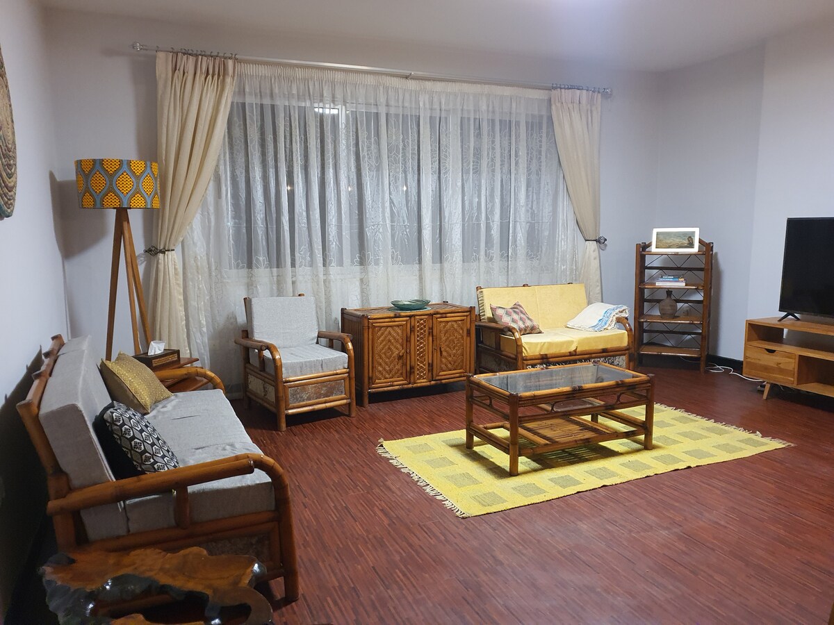 A spacious living area presents a combination of wooden furniture including a coffee table and two armchairs, complemented by a sofa with cushions. A vibrant yellow area rug enhances the space, while large windows are adorned with sheer curtains, allowing natural light to flow in.